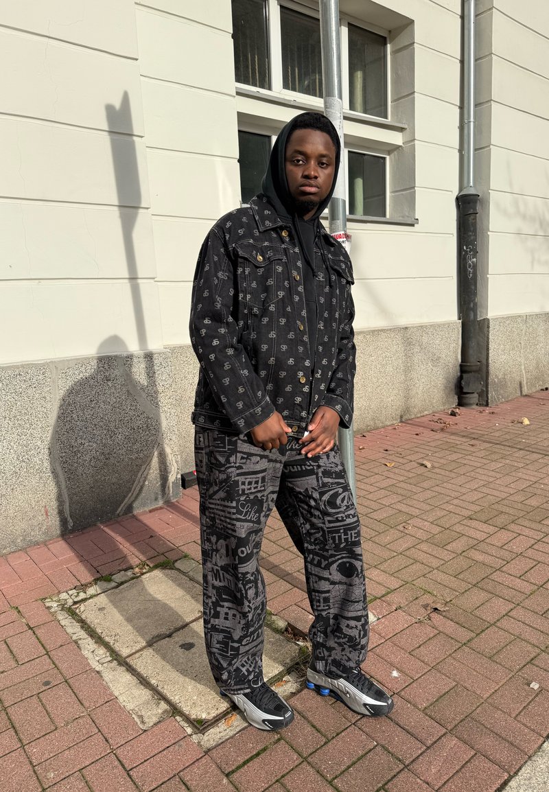 Chaqueta de mezclilla negra con logotipos sutiles, combinada con pantalones negros con estampado. Ambos tienen un ajuste relajado, completando el look con zapatillas deportivas.