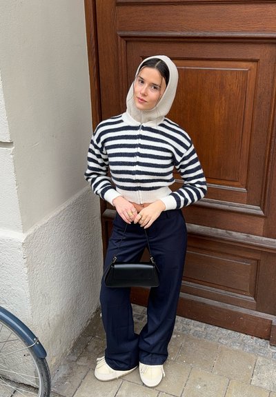 Cárdigan de punto a rayas en crema y azul marino, con cuello alto, combinado con pantalones anchos oscuros y un pequeño bolso negro. Calzado casual completa el look.