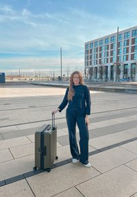 Mørkegrønn koffert med hardt skall og ribbet tekstur, utstyrt med uttrekkbart håndtak og fire hjul, plassert på en fortau nær en bygning.