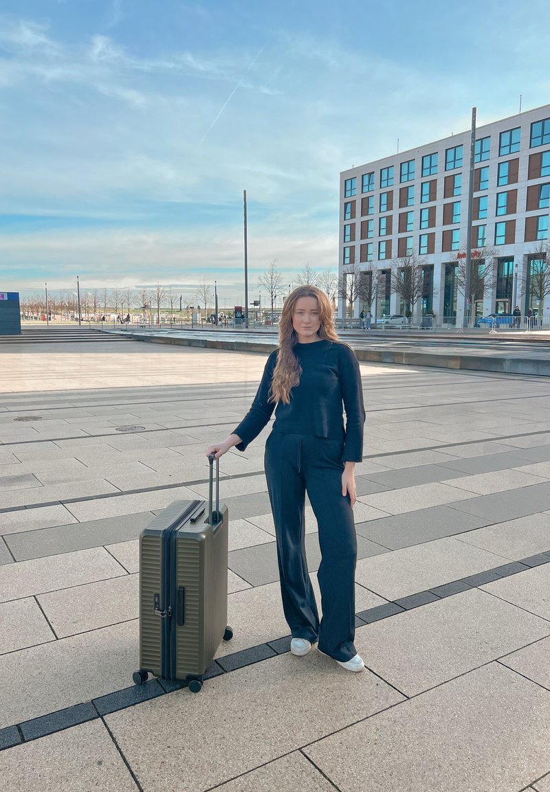 Valise rigide vert foncé à texture nervurée, poignée rétractable et quatre roues, posée sur un trottoir près d'un bâtiment.
