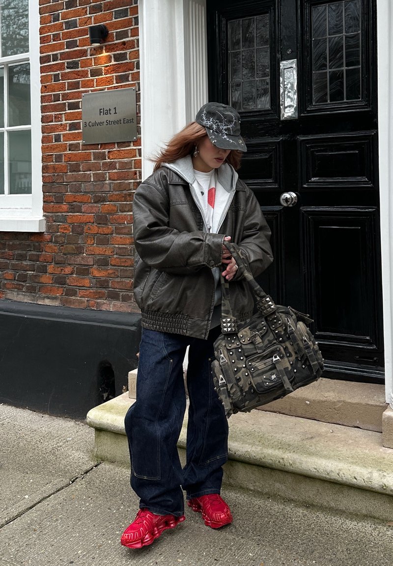 Personne portant une veste en cuir foncé, une casquette noire, des baskets rouges et un jean ample, debout près de l'entrée d'un bâtiment en briques, tenant un sac à motifs camouflage avec des clous.