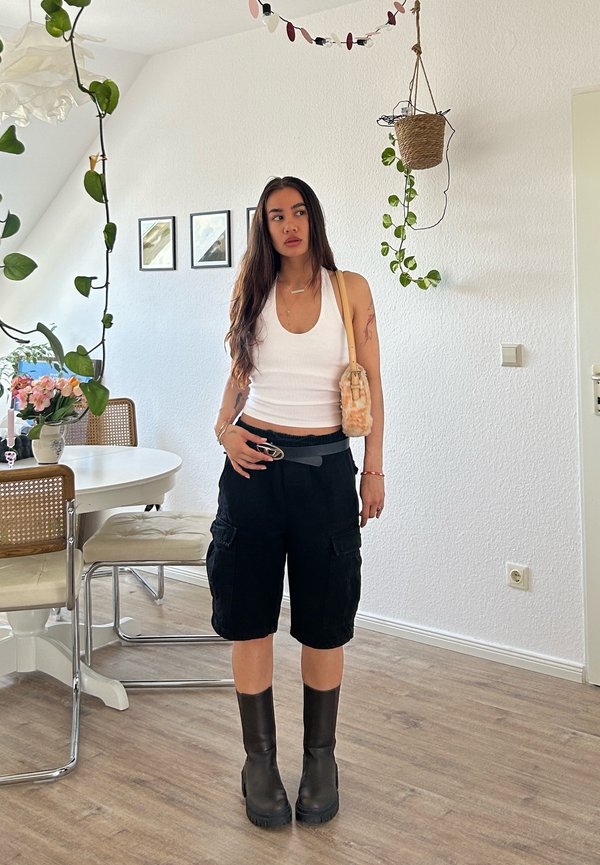 Kvinde med langt mørkt hår iført hvid tanktop, sorte cargoshorts, sorte støvler og bærer en lille beige skuldertaske i et lyst rum.