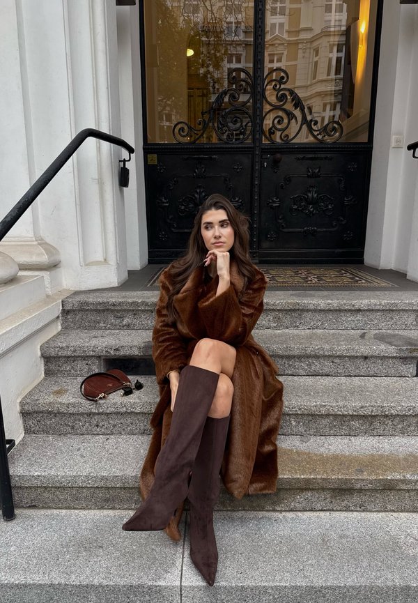 Cappotto di pelliccia sintetica marrone, stivali marroni in suede fino alla coscia con punte appuntite, seduta su gradini di pietra davanti a doppie porte nere ornate.