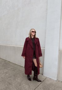 Burgundský trenčkot s dvojradovým zapínaním, doplnený priliehavým hnedým topom a mini sukňou. Hnedé čižmy ku kolenám a malá kabelka.