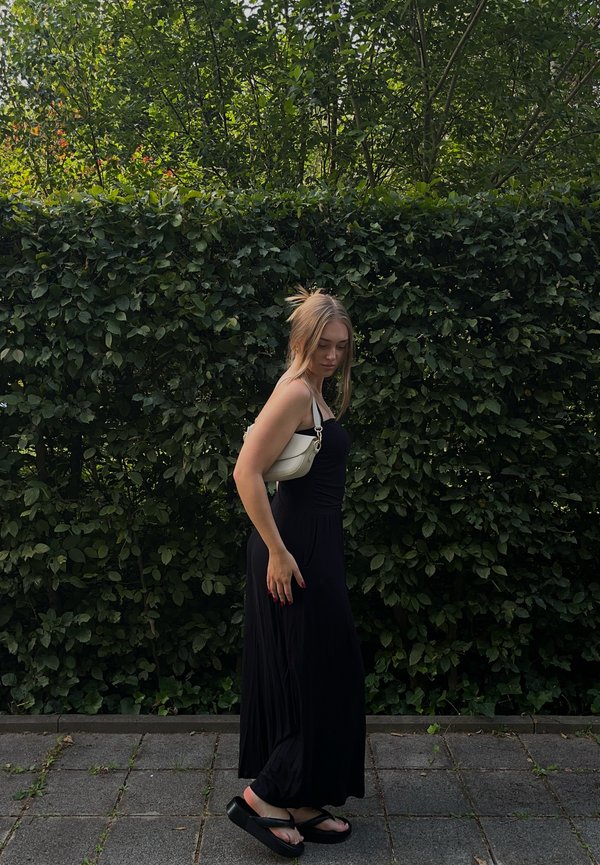 Black maxi dress crafted from a soft fabric; white crossbody bag with silver hardware accents. The background showcases lush greenery and a paved surface.