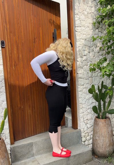 Persona con cabello rubio largo que lleva ropa blanca y negra y zapatos rojos, parada en escalones frente a una puerta de madera junto a una pared de piedra con plantas.