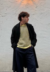 Homme aux cheveux bouclés portant un pull rayé jaune et marron, un manteau noir, un pantalon foncé et des écouteurs sans fil blancs, debout contre un mur texturé.