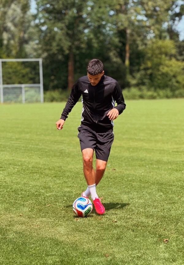 Jeune homme en tenue de sport noire dribblant un ballon de football coloré sur un terrain en herbe avec des buts et des arbres en arrière-plan.