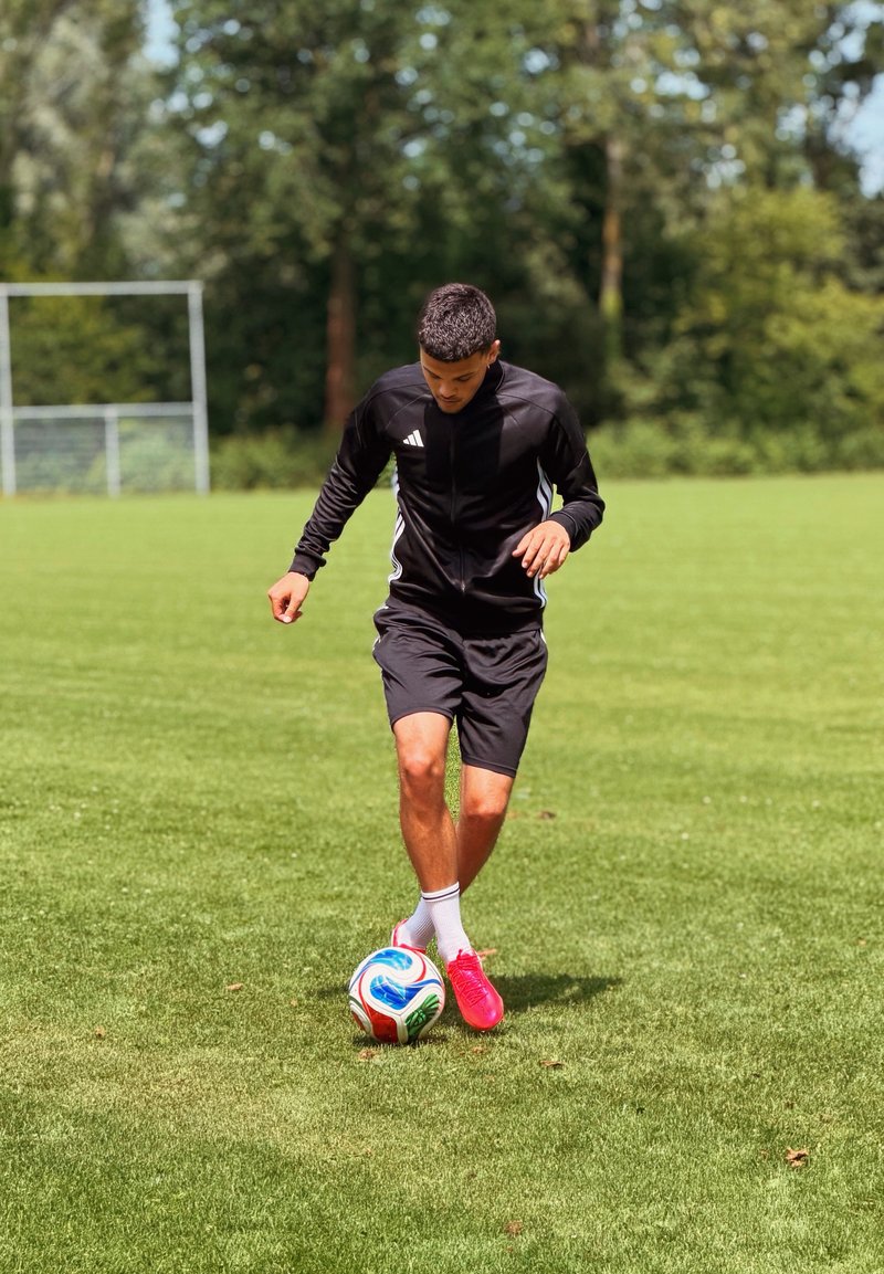 Jeune homme en tenue de sport noire dribblant un ballon de football coloré sur un terrain herbeux avec des buts et des arbres en arrière-plan.