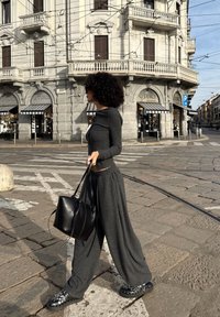 Maglione grigio scuro a maniche lunghe e pantaloni larghi abbinati, abbinati a una borsa nera in tessuto testurizzato e calzature nere con motivo, sullo sfondo urbano.