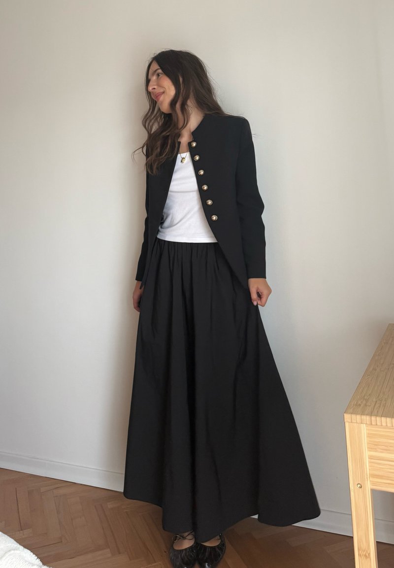 Schwarzer Blazer mit goldenen Knöpfen, weißes Oberteil und schwarzer Maxiröcke. Glatter Stoff, taillierte Passform, ausgestellt Rock und flache Schuhe runden das Outfit ab.