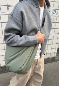 Hellgrüne, gepolsterte Umhängetasche mit einer geschwungenen Form, die crossbody getragen wird. Im Hintergrund sind ein grauer Reißverschluss-Hoodie und beigen Hosen sichtbar.