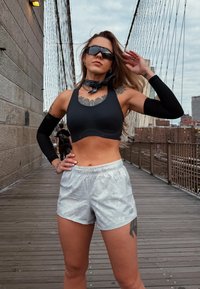 Čierna športová podprsenka, lesklé strieborné šortky s jemným vzorom, čierne rukávce a čierny bandana. Modelka pózuje na drevenom moste.