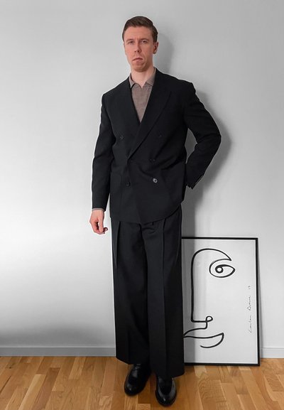 Hombre con blazer negro cruzado, pantalones negros anchos y zapatos negros, de pie junto a un retrato minimalista en líneas de un rostro enmarcado contra una pared blanca.