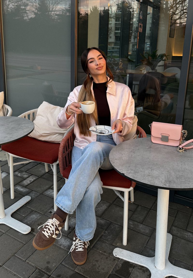 Femme assise à une table de café en plein air, tenant une tasse et une soucoupe, portant une veste rose clair, un haut noir et un jean bleu, avec un sac rose à proximité.
