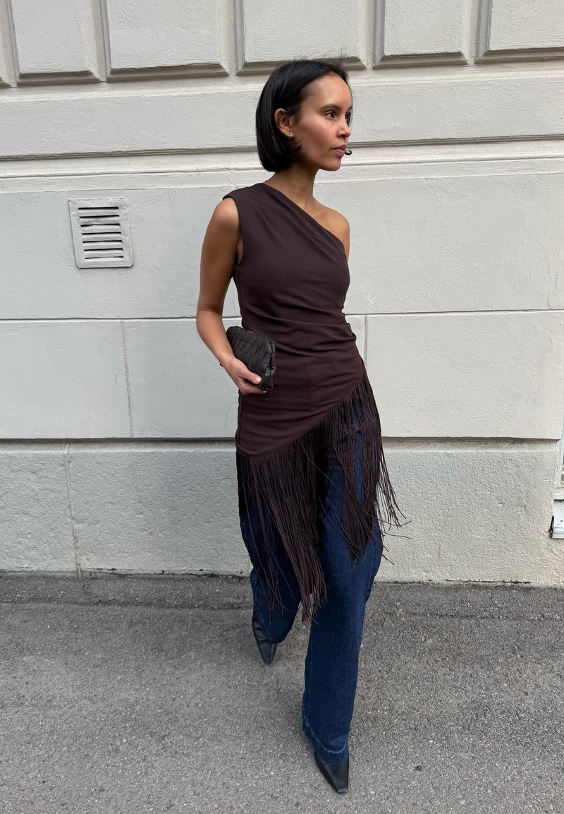 Femme aux cheveux courts portant un haut noir asymétrique à franges, jeans foncés, chaussures noires pointues, tenant une petite pochette noire tissée, marchant en extérieur.