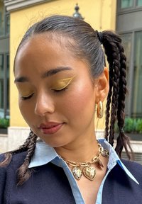 Gul hjärtformat smycke och en kedja pryder halslinjen; gul ögonskugga med djärv eyeliner kontrasterar mot brun hud och mörkt hår med flätor.