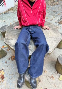 Červená větrovka s bílými pruhy, tmavě modré džíny se širokými nohavicemi a černé boty, sedící na dřevěné lavici v přírodním venkovním prostředí.