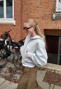 Hellgrauer Kapuzenpullover mit Reißverschluss, lockerer Passform und großen Taschen, kombiniert mit einem langen braunen Rock. Im Hintergrund sind Fahrräder und eine Backsteinmauer zu sehen.