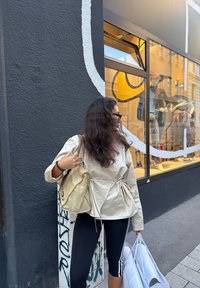 Beige letvægtsjakke med snøre i taljen, sorte leggings med hvide striber, holder en guldfarvet håndtaske og hvide indkøbsposer. Urban baggrund.