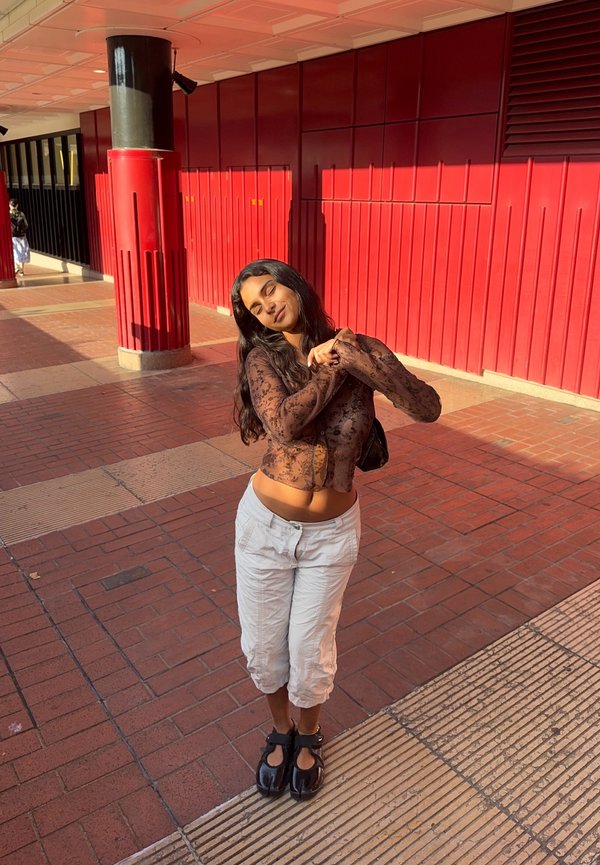 Mujer con pantalones blancos y blusa marrón translúcida, de pie con las manos juntas, ojos cerrados, en un pasillo iluminado por el sol con paredes y columnas rojas.