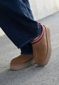 Barna szarvasbőr bebújós cipők vastag talppal és mintás, piros díszítéssel a boka körül, sötétkék farmernadrággal viselve.