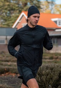 Fekete futó dzseki cipzárral és kiegészítőkkel, fekete kesztyűvel és sorttal párosítva. Az anyag könnyűnek és rugalmasnak tűnik.