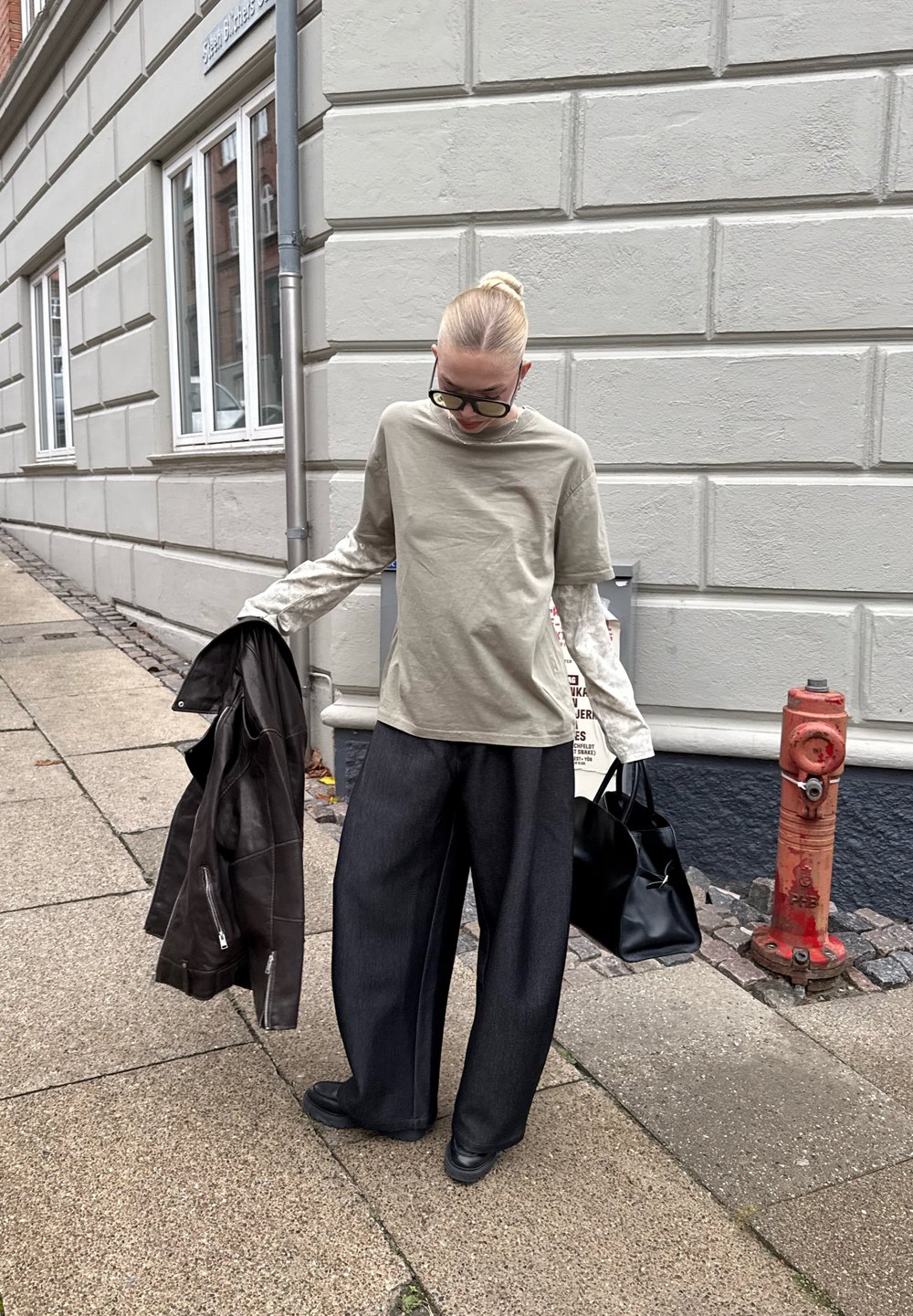 Lichtolijfgroene T-shirt, gecombineerd met witte lange mouwen, en brede donkergrijze broek met een zwarte handtas. Bruine leren jas los over de schouder gehangen.