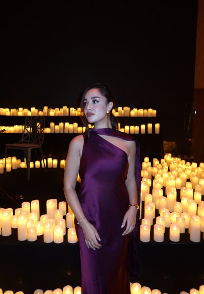 Mujer con un vestido de satén morado de pie entre numerosas velas encendidas colocadas sobre superficies negras en una habitación con poca luz, con una silla moderna al fondo.