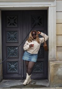 Giacca corta beige, minigonna di denim, borsa marrone, occhiali da sole scuri. Stivali da cowboy bianchi, con una tazza di caffè in mano, appoggiata a una porta.