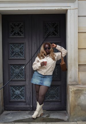Chaqueta corta beige, minifalda vaquera, bolso marrón, gafas de sol oscuras. Botas de vaquero blancas, sosteniendo una taza de café, apoyada contra una puerta.