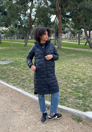 Abrigo de plumas negro con costuras onduladas y diseño con capucha. Combinado con jeans azules y zapatillas negras. Entorno al aire libre con hierba y árboles.