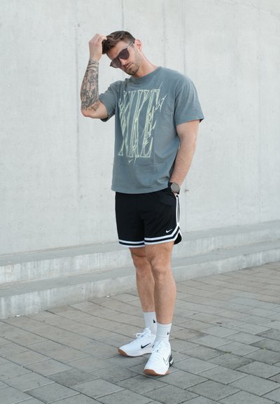 Hombre con gafas de sol, camiseta gris de Nike, pantalones cortos negros de Nike y zapatillas blancas, de pie sobre una acera pavimentada frente a un muro de hormigón.