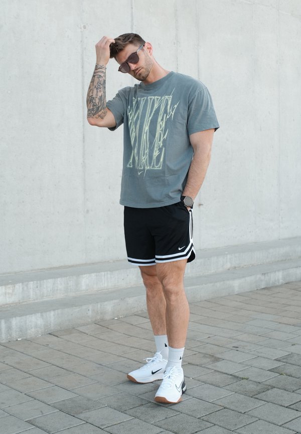 Muž nosící sluneční brýle, šedé tričko Nike, černé kraťasy Nike a bílé tenisky, stojící na dlážděném chodníku u betonové zdi.