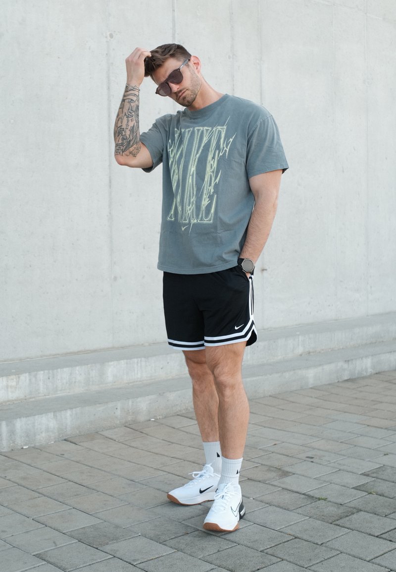 Homme portant des lunettes de soleil, un t-shirt gris Nike, un short noir Nike et des baskets blanches, debout sur un trottoir pavé contre un mur en béton.