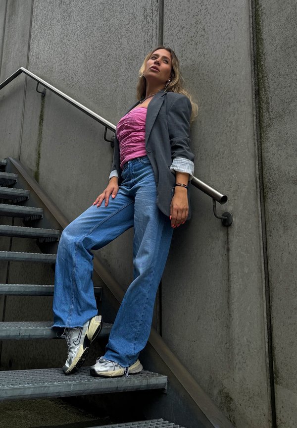 Grå blazer over en lyserød krøllet top, parret med løse blå jeans og metalliske sølv sneakers. Model læner sig op ad en grå væg.