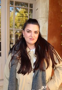 Beżowy trencz z złotymi elementami, nałożony na biały top, uzupełniony szarym szalikiem z wzorzystą krawędzią. Długie ciemne włosy.