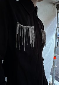 Musta nappikauluspaita, jossa on kaksi taskua, koristeltu pitkällä, hopeisella kimalteella, luoden tekstuurisen ja dynaamisen visuaalisen vaikutelman.