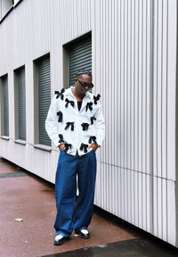 Hvit skjorte med svarte sløyfedetaljer, kombinert med løse dongeribukser og svart-hvite sko. Bakgrunnen har vertikal panelvegg.