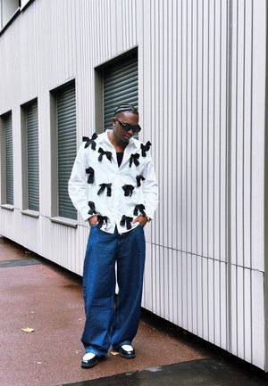 Camisa blanca de botones con detalles de lazo negro, combinada con pantalones vaqueros holgados y zapatos en blanco y negro. El fondo presenta paneles verticales.