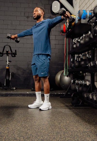 Hombre de pie en el gimnasio levantando mancuernas lateralmente, vistiendo ropa deportiva azul y zapatillas blancas cerca de equipos de ejercicio.