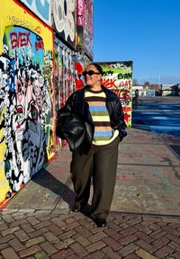 Giacca in pelle nera, maglione a righe sfocate verde, marrone e blu, pantaloni larghi marroni e borsa texturizzata davanti a un muro di graffiti colorati.