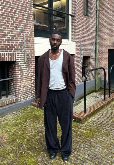 Blazer oversized de color marrón oscuro sobre una camiseta gris clara, combinado con pantalones anchos negros. Situado frente a una pared de ladrillo y un suelo pavimentado.