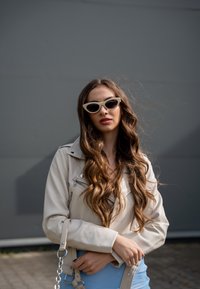 Κρέμ δερμάτινο μπουφάν μοτοσικλετιστή με φερμουάρ, συνδυασμένο με ελαφρύ μπλε φούστα. Το μοντέλο φοράει λευκά γυαλιά ηλίου cat-eye και έχει μακριά, κυματιστά μαλλιά.