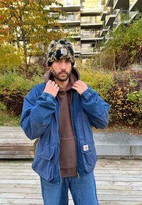 Denimjacke aus blauer Baumwolle, darunter eine beige Fleecekapuzenjacke und eine Mütze in Schwarz- und Olivgrünmuster. Hände sichtbar, die Ringe zeigen.