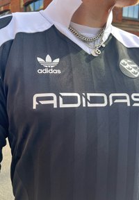 Μαύρη φανέλα Adidas με λευκό γιακά και λεπτομέρειες, που διαθέτει οριζόντιες ρίγες. Διακοσμημένη με το λογότυπο και το όνομα της μάρκας Adidas σε ασημί χρώμα.