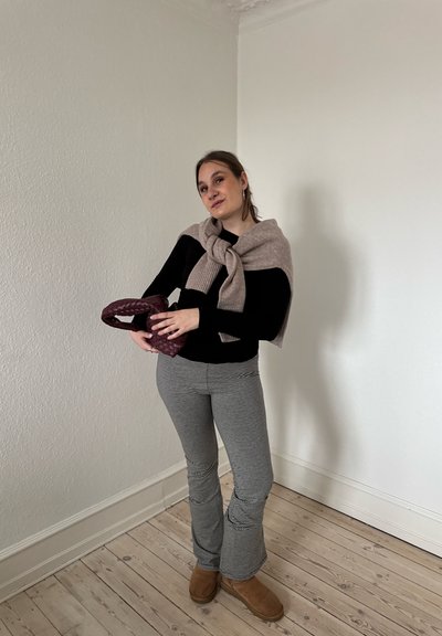 Mujer con suéter beige atado alrededor de los hombros, camiseta negra de manga larga, pantalones a rayas y botas marrones sosteniendo un bolso tejido color granate en una habitación minimalista.