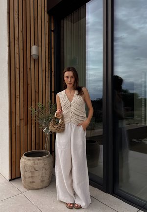 Chaleco de ganchillo color beige con escote en V y botones al frente, combinado con pantalones de lino blancos holgados. Sosteniendo un bolso de mimbre, de pie junto a una ventana.
