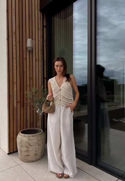 Chaleco de ganchillo color beige con escote en V y botones al frente, combinado con pantalones de lino blancos holgados. Sosteniendo un bolso de mimbre, de pie junto a una ventana.