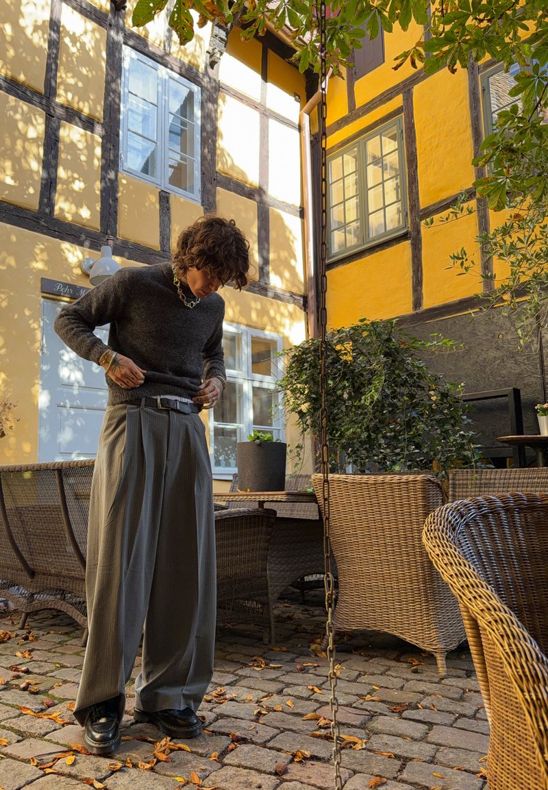 Grau gestrickter Pullover, gefaltete graue Hose und schwarze Schuhe. Außenhof mit Steinboden, Rattanmöbeln und gelben Wänden.